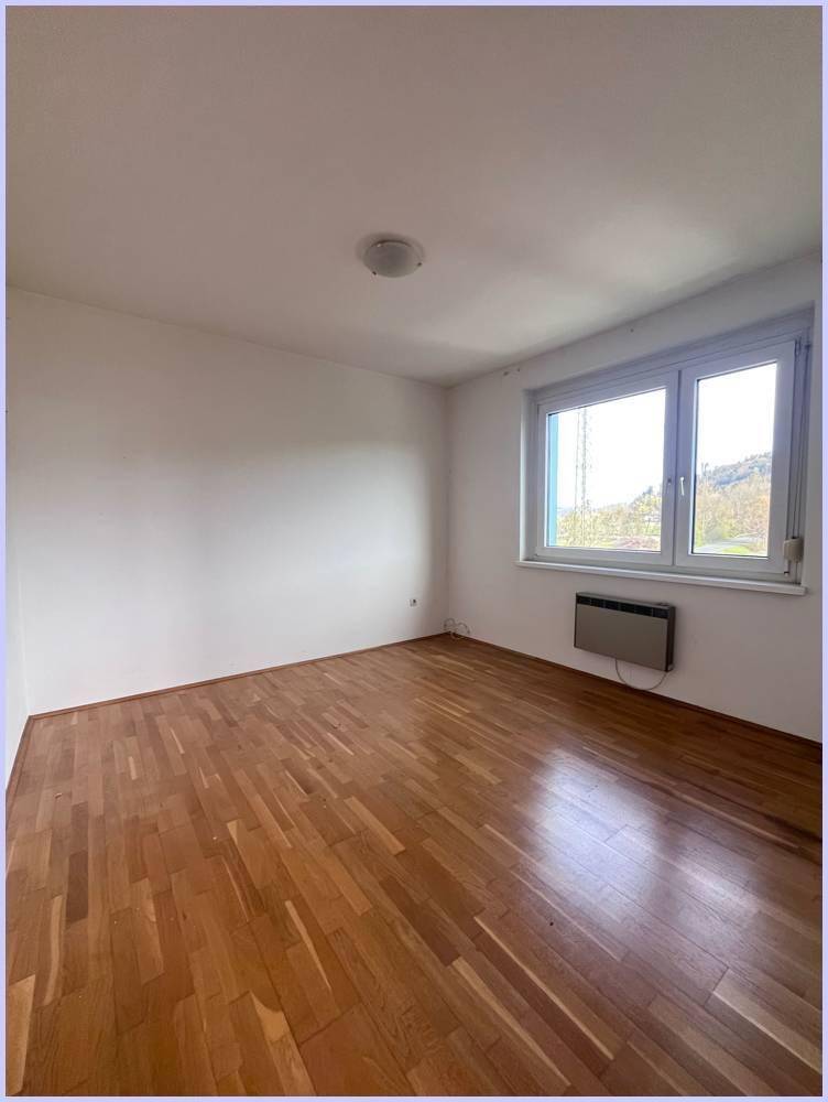 Leeres Zimmer mit Holzboden, großem Fenster und einem Heizkörper an der Wand.