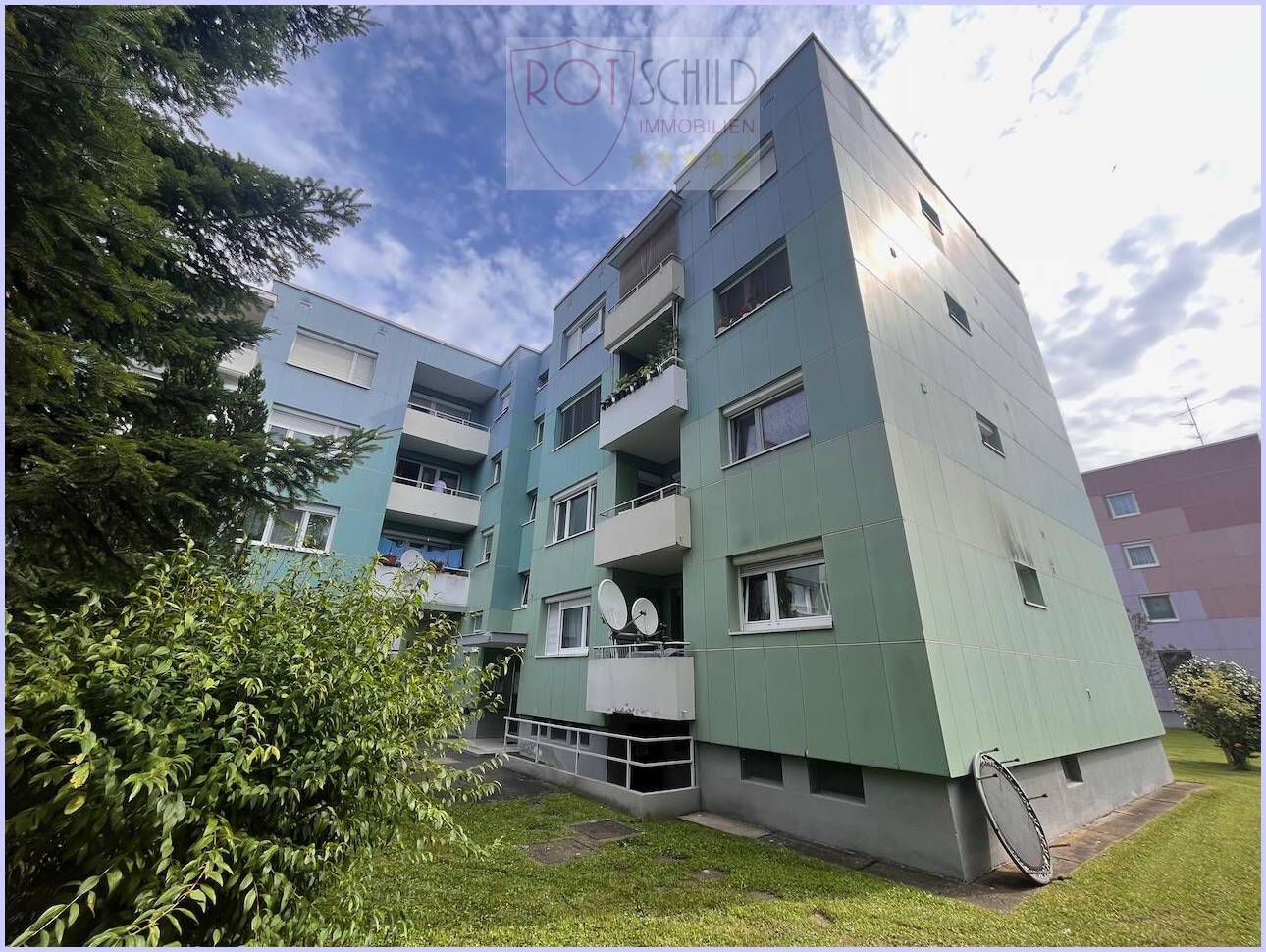 Mehrstöckiges Wohngebäude mit Balkonen und grüner Fassade, umgeben von Bäumen.