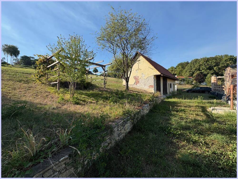 Gartenbereich mit einem Gebäude, einer Steinmauer und einer Holzpergola, umgeben von ländlicher Natur.