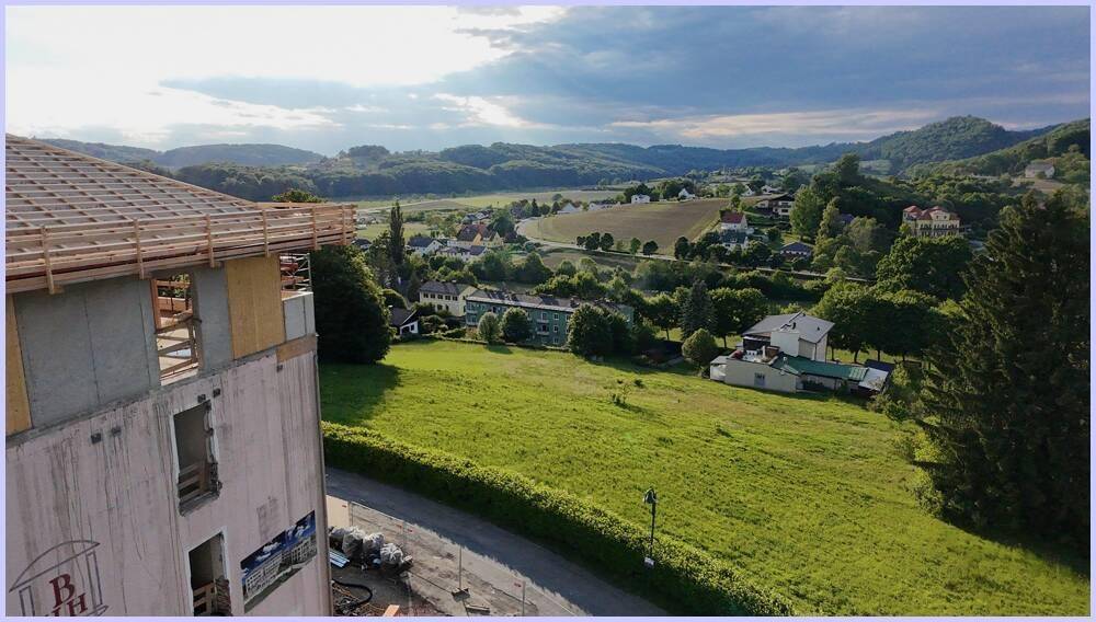 Gebäude im Bau mit Blick auf eine weite grüne Landschaft, ein Dorf und bewaldete Hügel unter bewölktem Himmel.