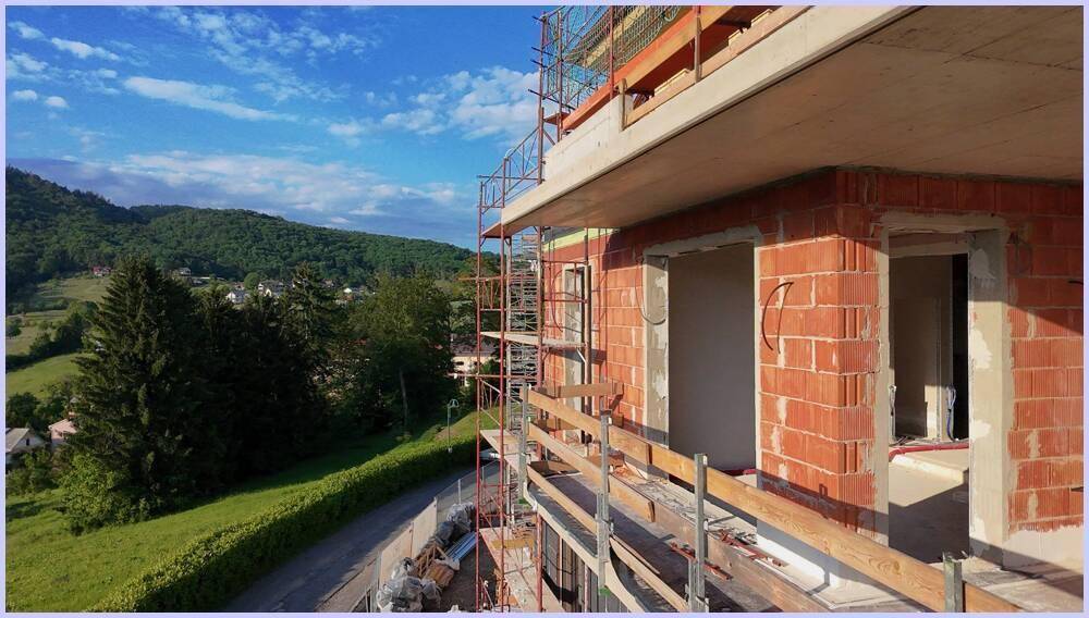 Nahaufnahme eines im Bau befindlichen Gebäudes mit Ziegelwänden und Gerüst vor grüner Landschaft.