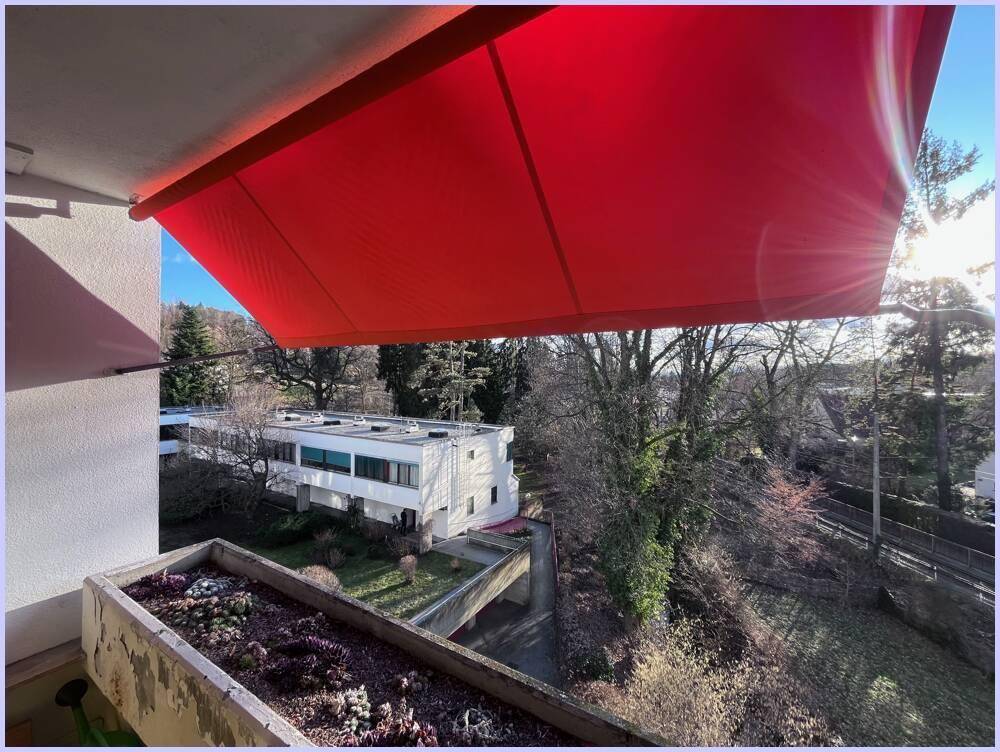 Balkon mit ausgefahrener roter Markise und weitem Blick über die grüne Umgebung.