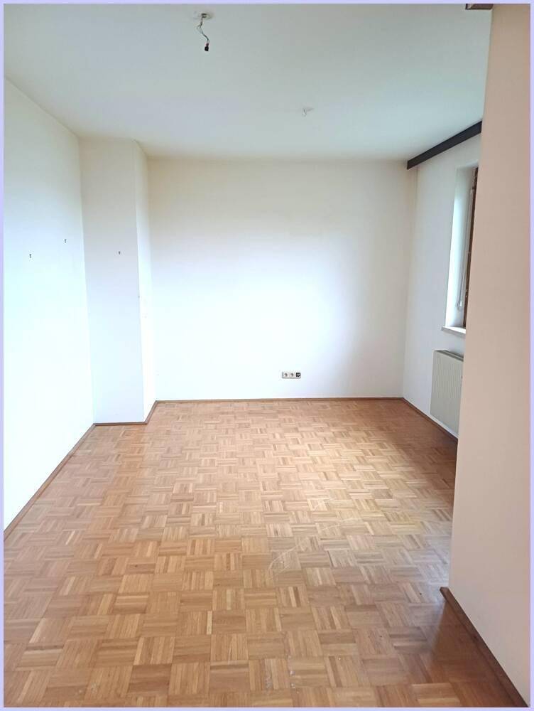Leeres Zimmer mit Parkettboden, weißen Wänden und einem Fenster mit Heizkörper.