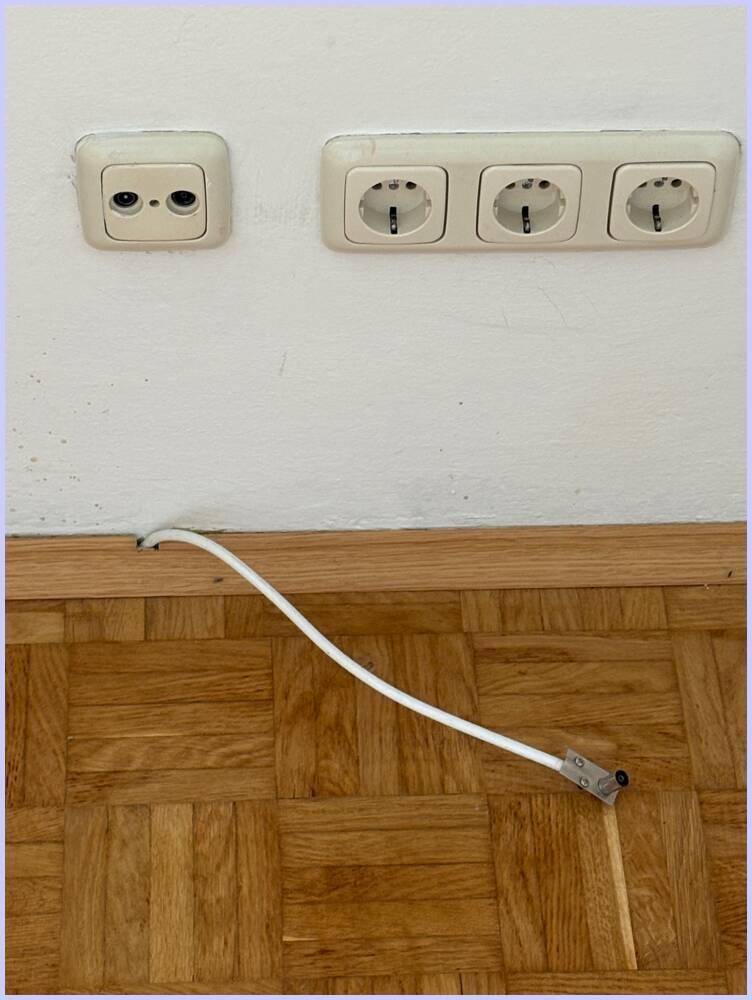 Mehrere Steckdosen und ein Kabelanschluss an einer weißen Wand über einem Parkettboden.