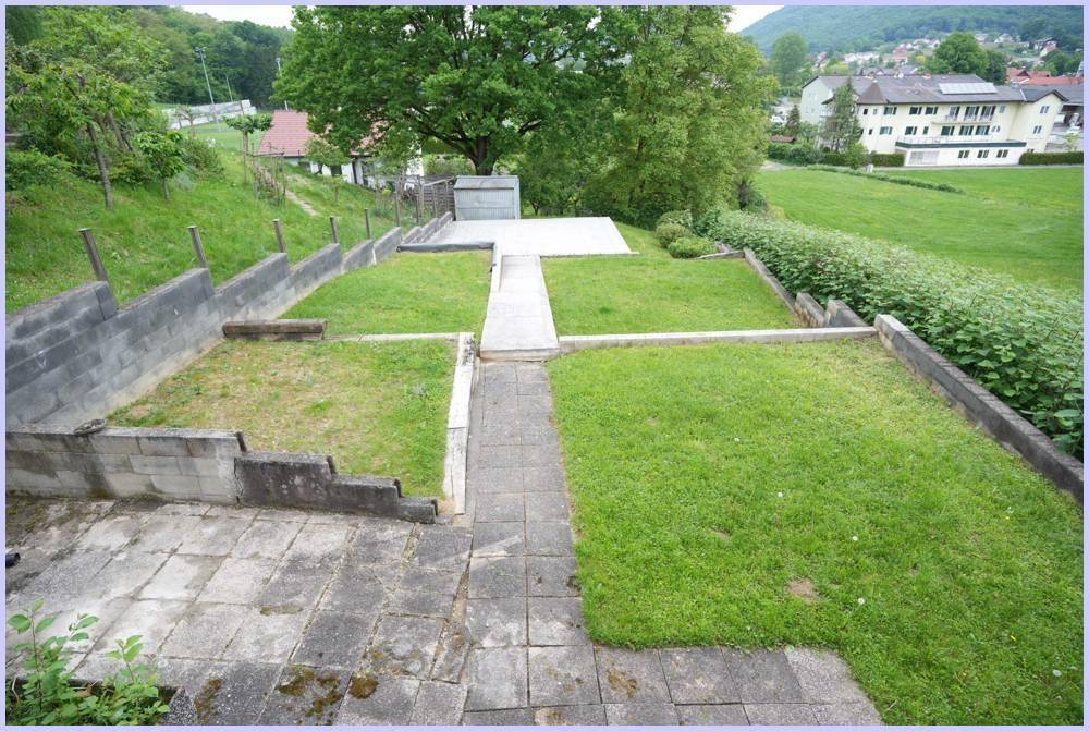 Gestufter Gartenbereich mit Rasenflächen und gepflasterten Wegen, umgeben von Betonmauern.