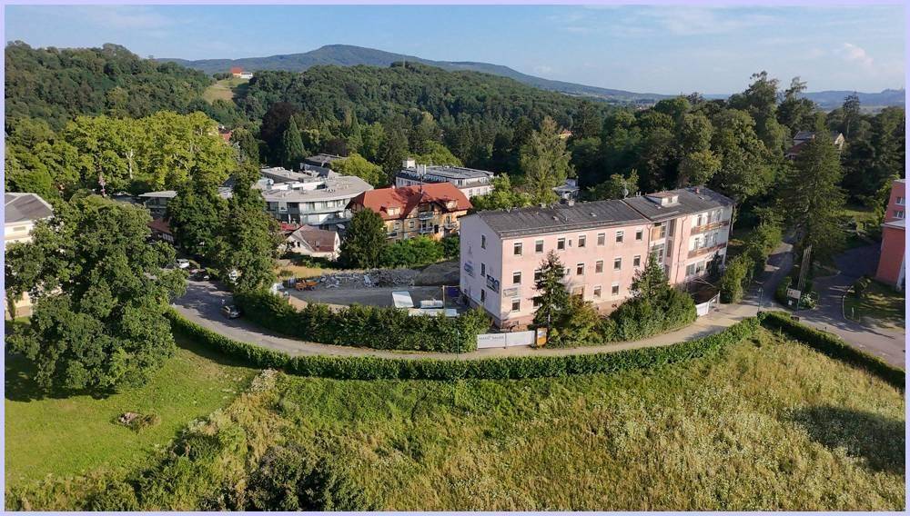 Luftaufnahme eines Gebäudekomplexes in einer grünen, hügeligen Landschaft mit umliegenden Bäumen und Wiesen.