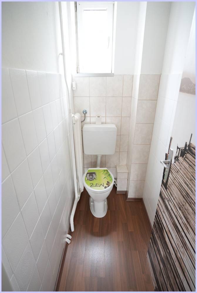 Kleines WC mit weißen Wandfliesen, Fenster und einem WC mit grünem Deckel.