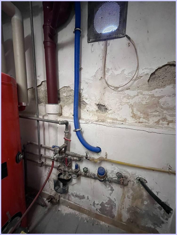 Detailansicht von Rohrleitungen und Ventilen an einer Wand mit abblätterndem Putz in einem Kellerraum.