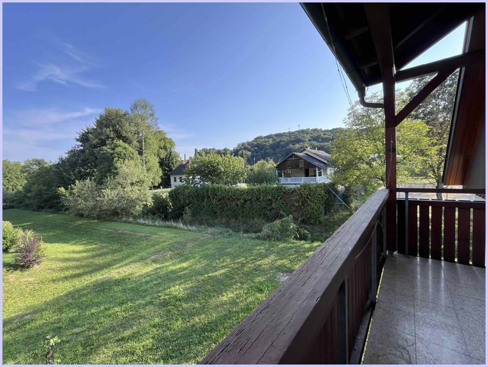 Sonniger Balkon mit Blick auf die grüne Hügellandschaft und ein charmantes Haus in der Ferne.
