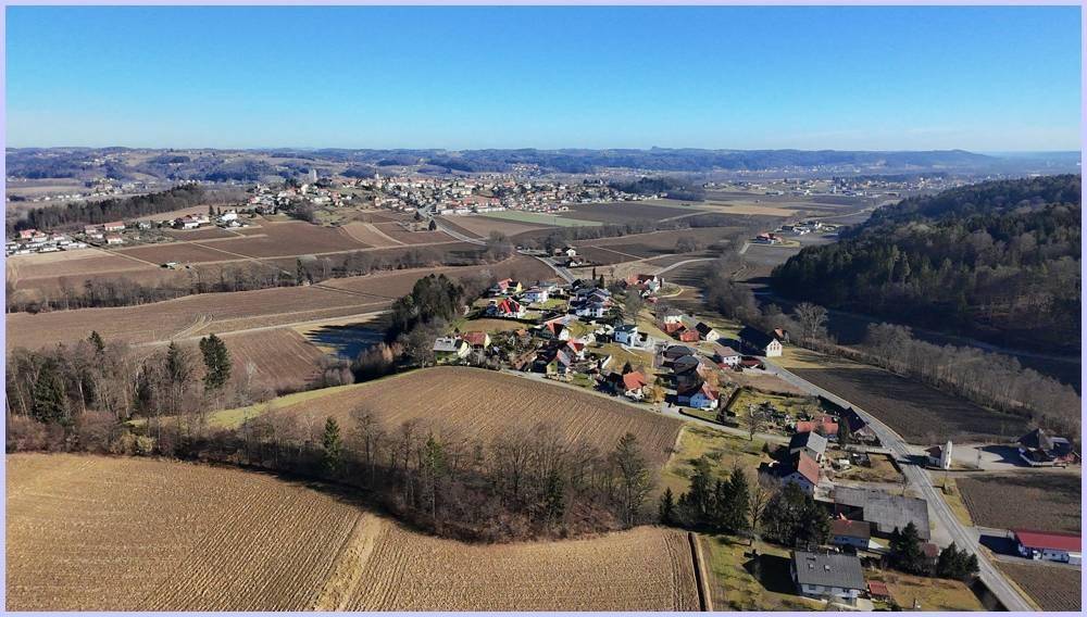 Weitläufige Luftaufnahme eines Tals mit einem Dorf, Feldern und einer Bergkette am Horizont.