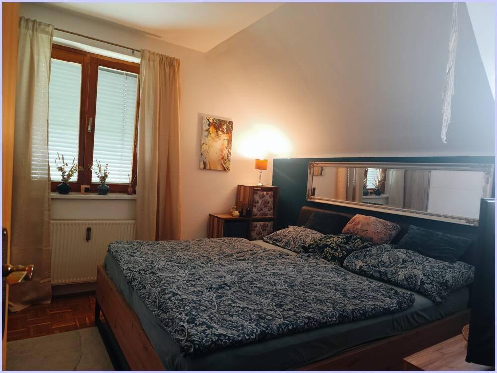 Schlafzimmer mit Doppelbett, Fenster und Dachschräge, beleuchtet von einer Lampe.