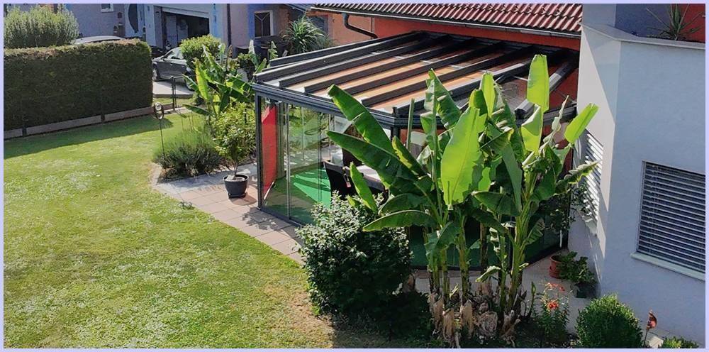 Gartenbereich mit Wintergarten und tropischen Pflanzen, umgeben von Rasenflächen.