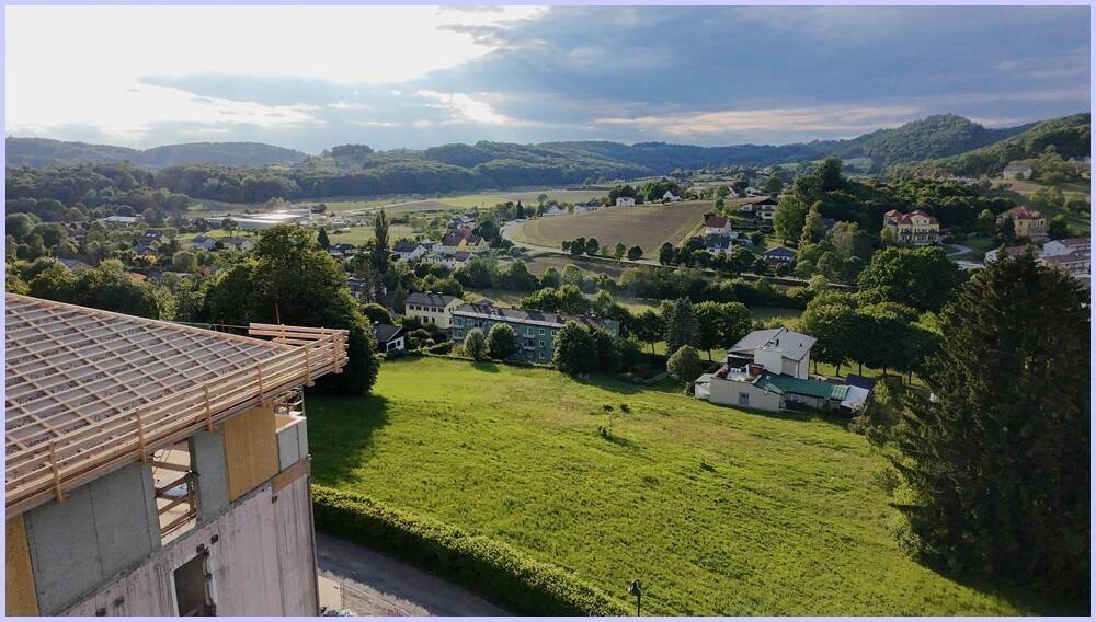 Gebäude im Bau mit weitem Blick über eine grüne Landschaft, ein Dorf und bewaldete Hügel unter bewölktem Himmel.