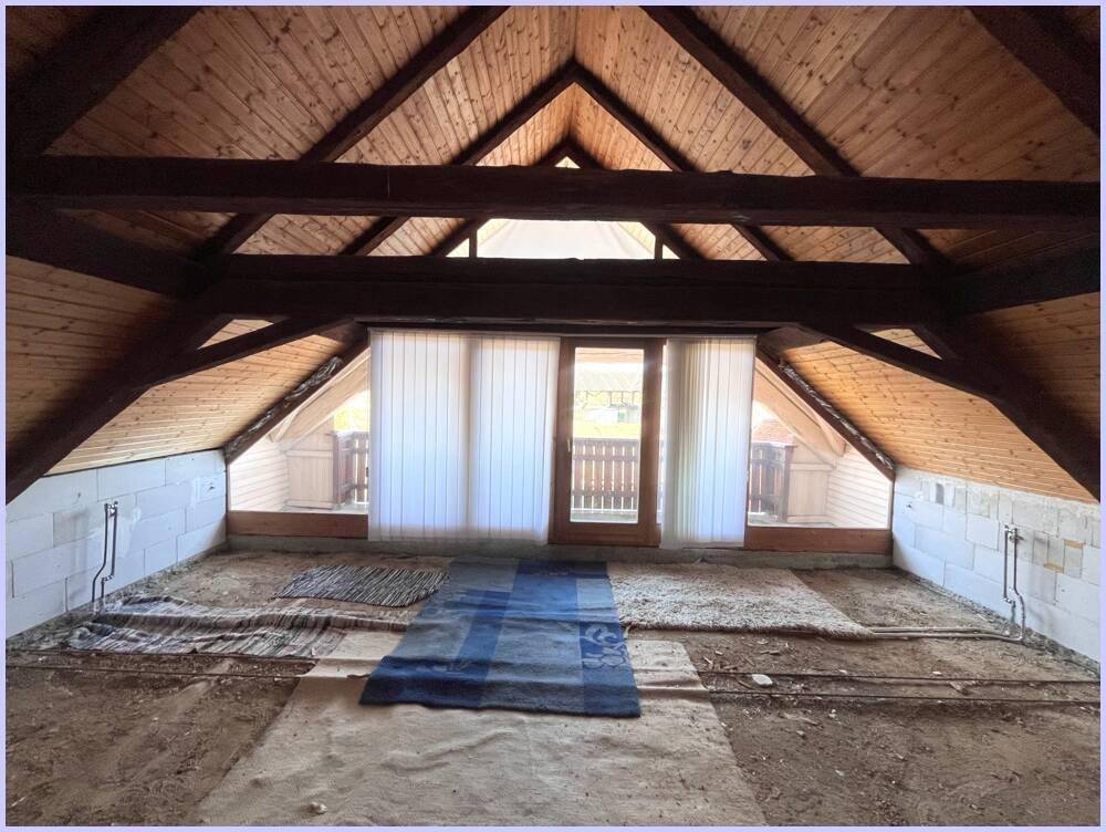 Unausgebauter Dachbodenbereich mit sichtbarer Holzkonstruktion und einem Fenster, das Tageslicht hereinlässt.