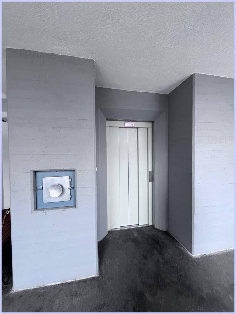 Heller Flur mit grauen Wänden, dunklem Boden und einem modernen Aufzug.