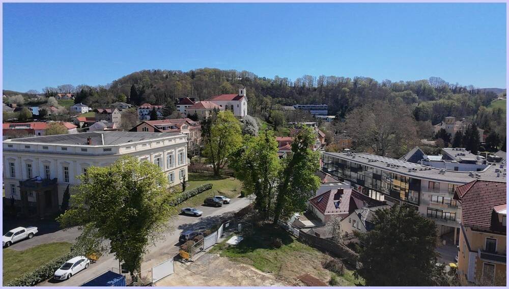 Luftaufnahme einer Stadtlandschaft mit verschiedenen Gebäudetypen und einer Kirche auf einem Hügel, umgeben von Natur.