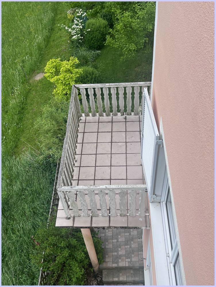 Balkon mit Fliesenboden und Geländer, von oben gesehen, umgeben von viel Grün.