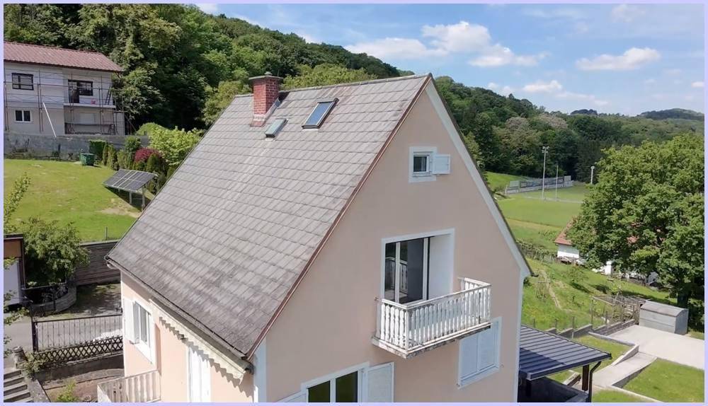 Ansicht des Hauses von oben mit Dach, Schornstein und Balkon in grüner Umgebung.