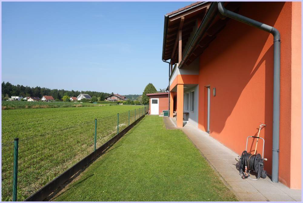 Seitenansicht des Hauses mit Rasenfläche, Zaun und weitem Blick auf grüne Felder.