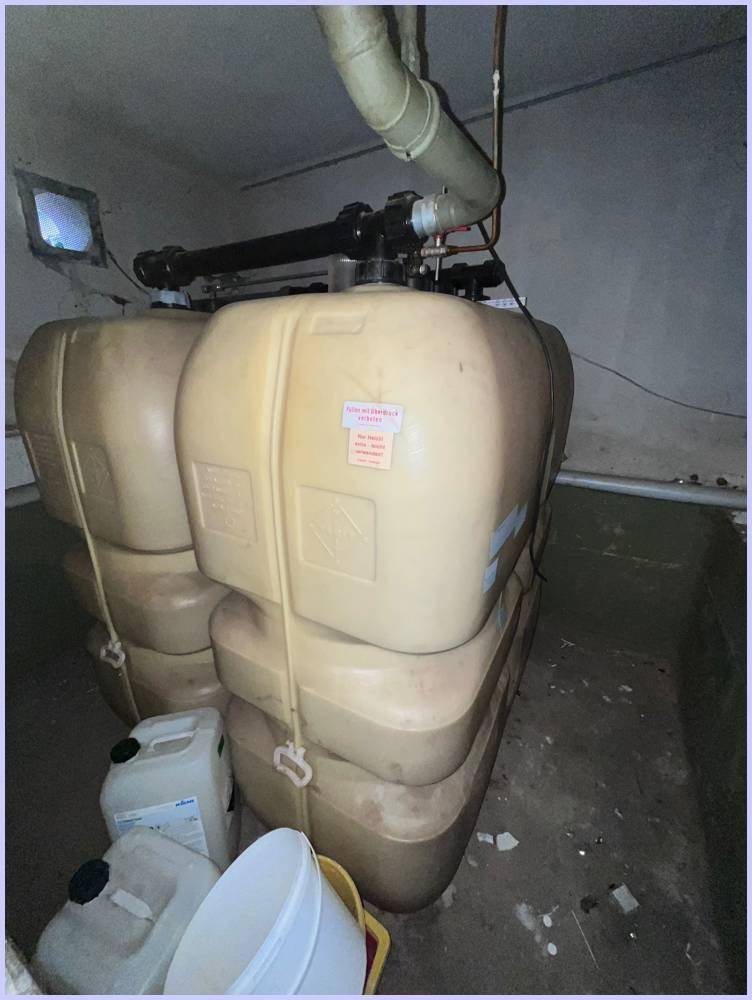 Mehrere große Kunststofftanks in einem Kellerraum, umgeben von Rohren und einer einfachen Beleuchtung.