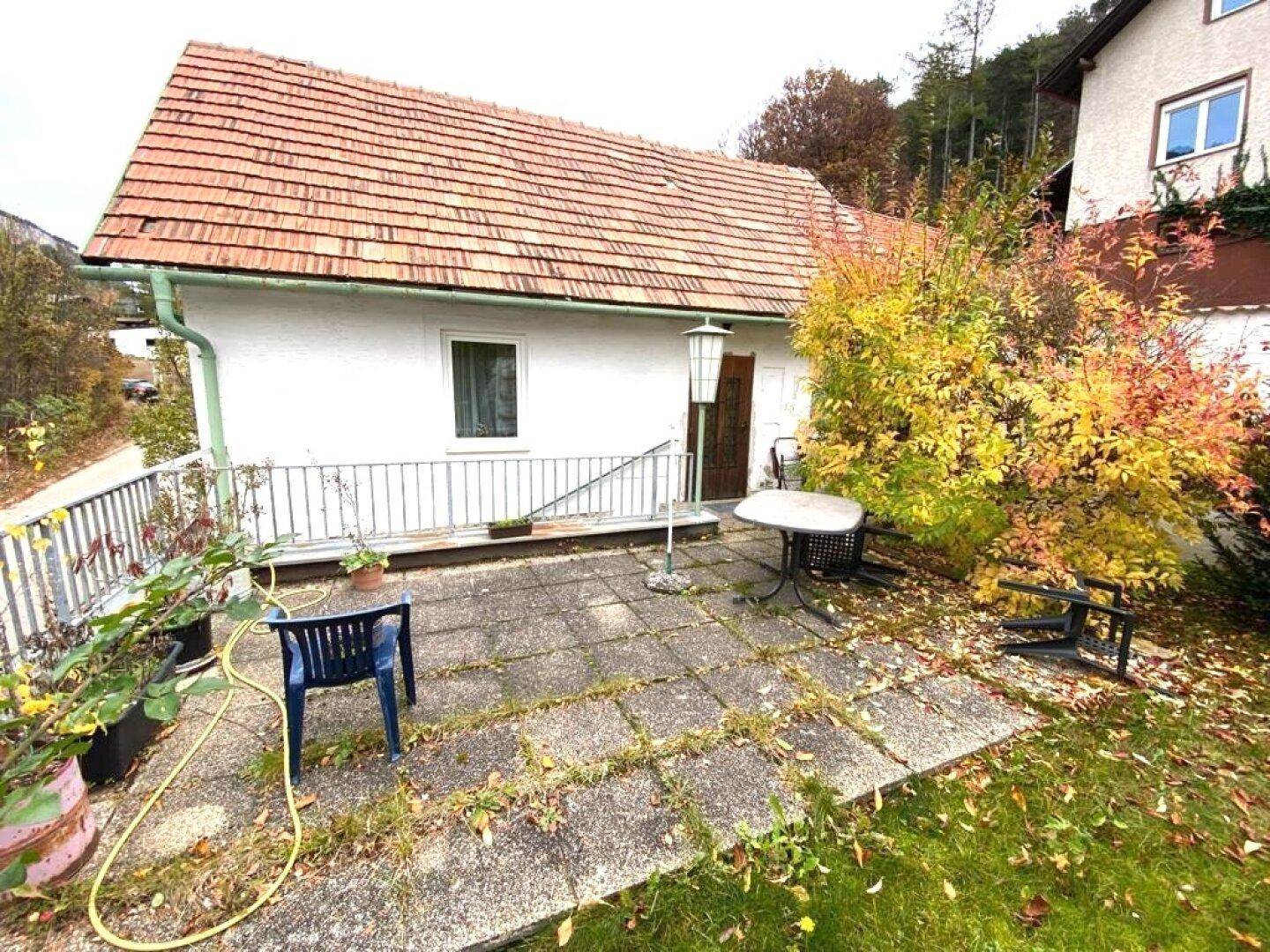 Einfache Terrasse mit Betonplatten und Gartenmöbeln, umgeben von herbstlichem Laub.