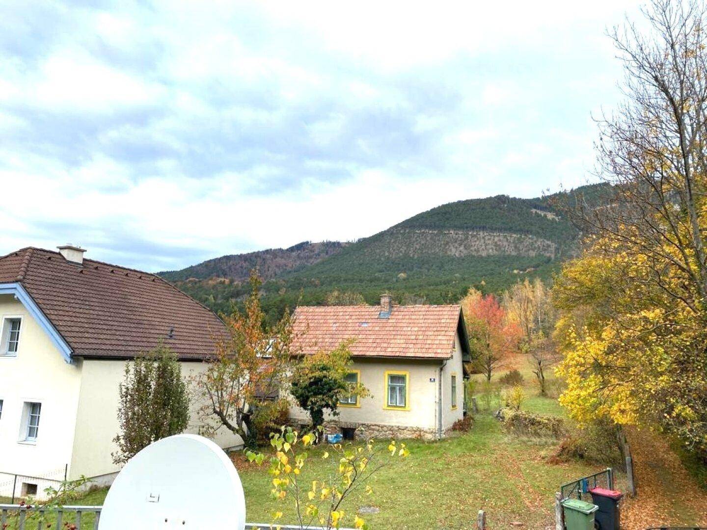 Herbstliche Landschaft mit zwei Häusern und bewaldeten Hügeln unter bewölktem Himmel.