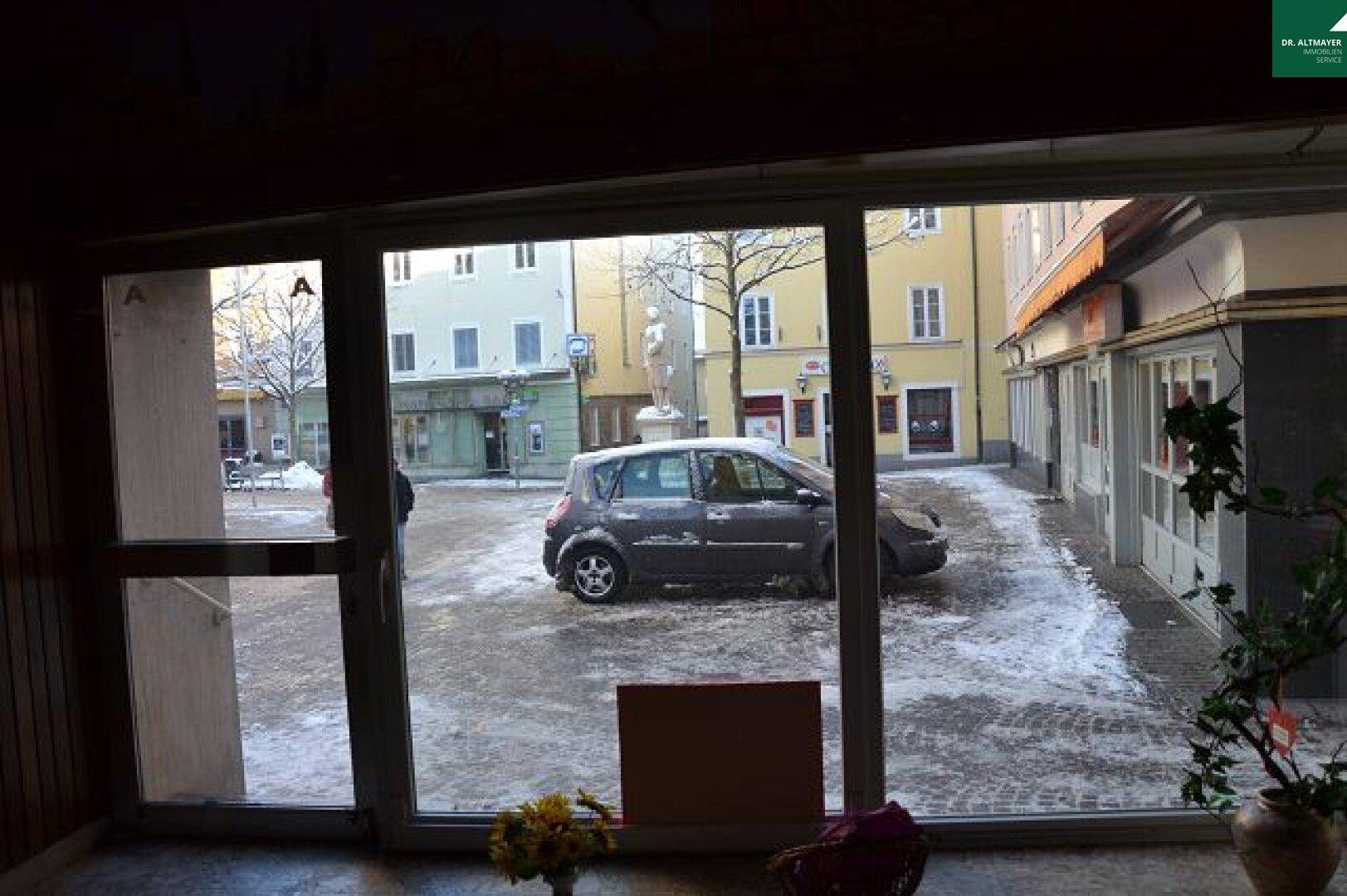 Blick aus einem Innenraum durch große Fenster auf einen verschneiten Stadtplatz mit parkendem Auto.