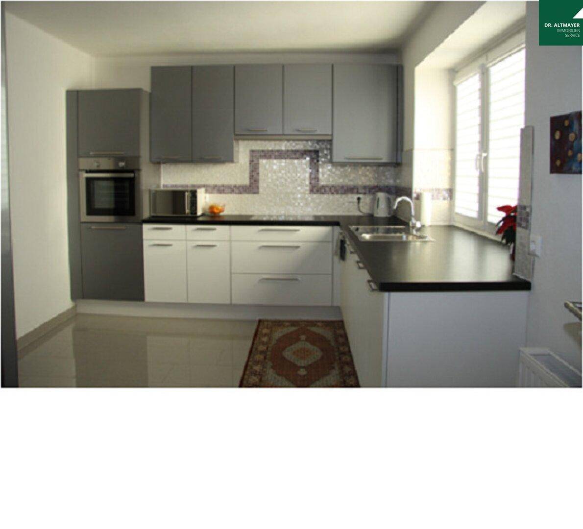 Moderne Küche mit grauen und weißen Schränken, dunkler Arbeitsplatte und Fenster für Tageslicht.