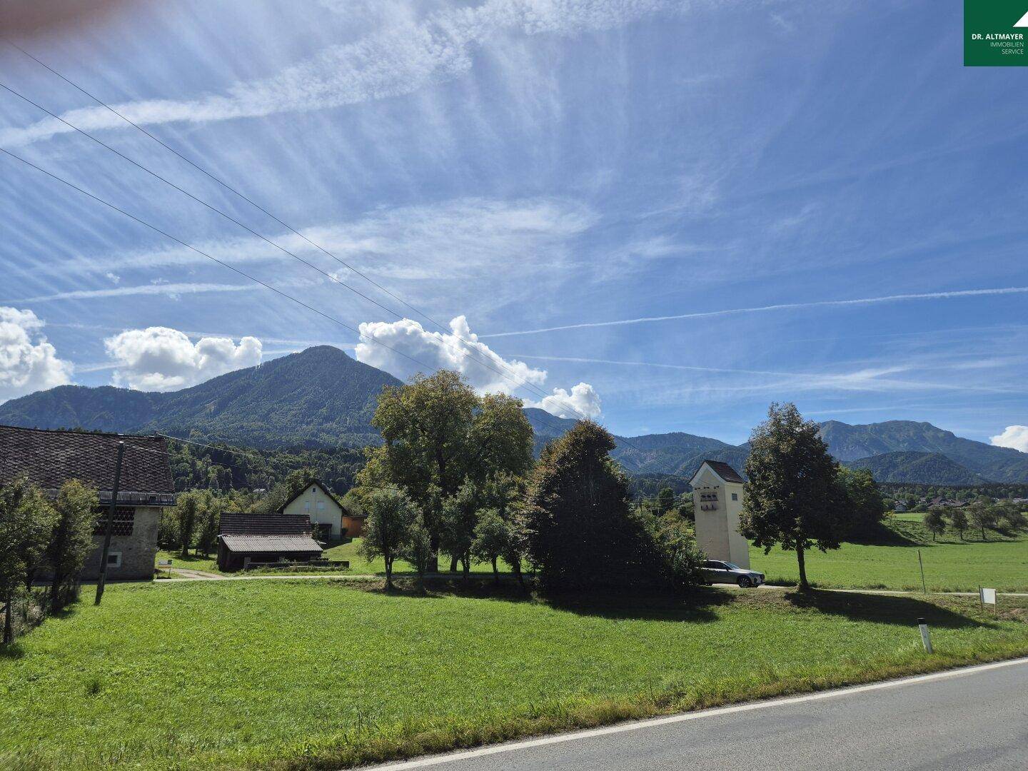 Weitläufige Landschaft mit grünen Wiesen, Bergen im Hintergrund und vereinzelten Häusern.