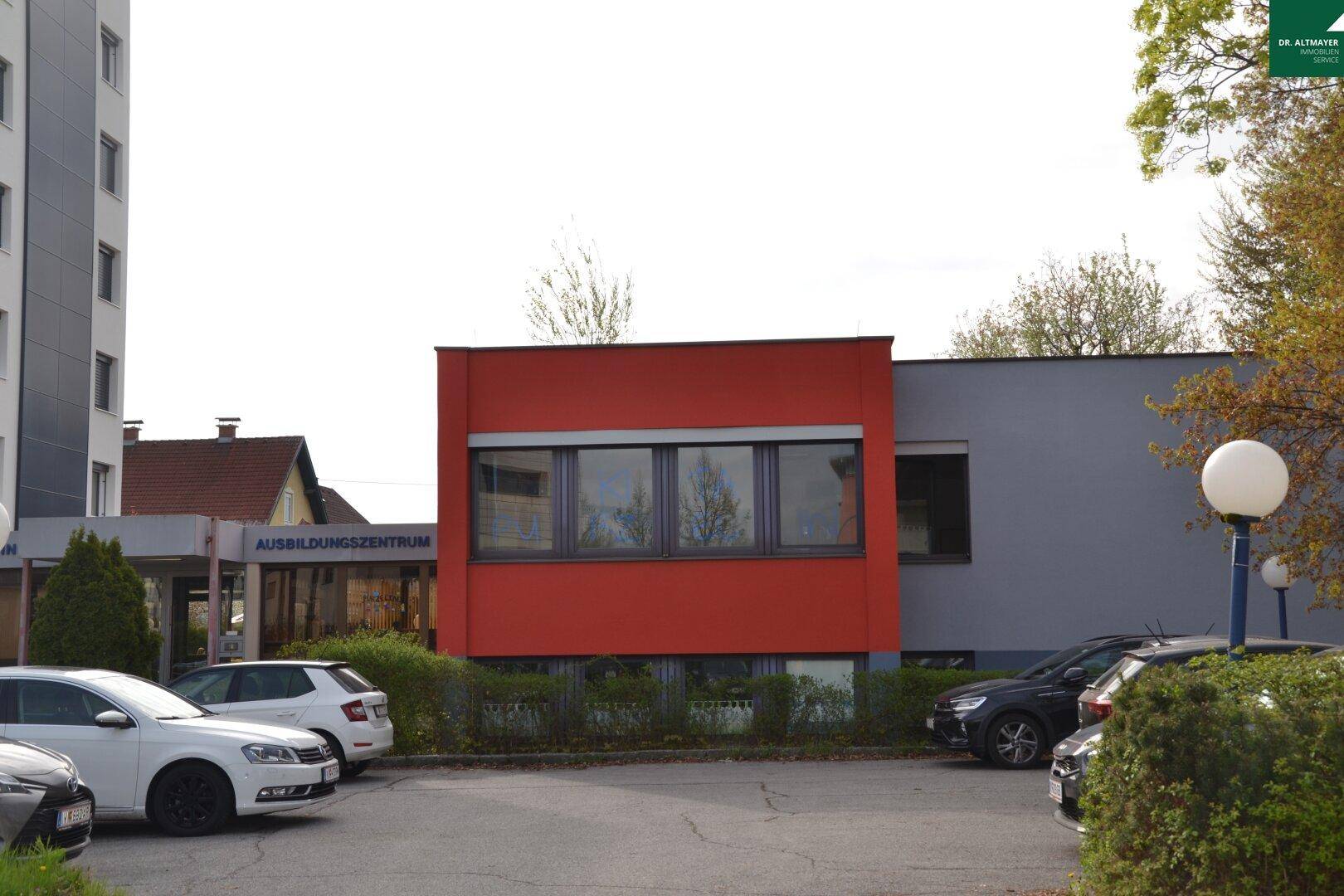 Gebäude mit markanter roter und grauer Fassade, daneben ein Parkplatz mit geparkten Autos.