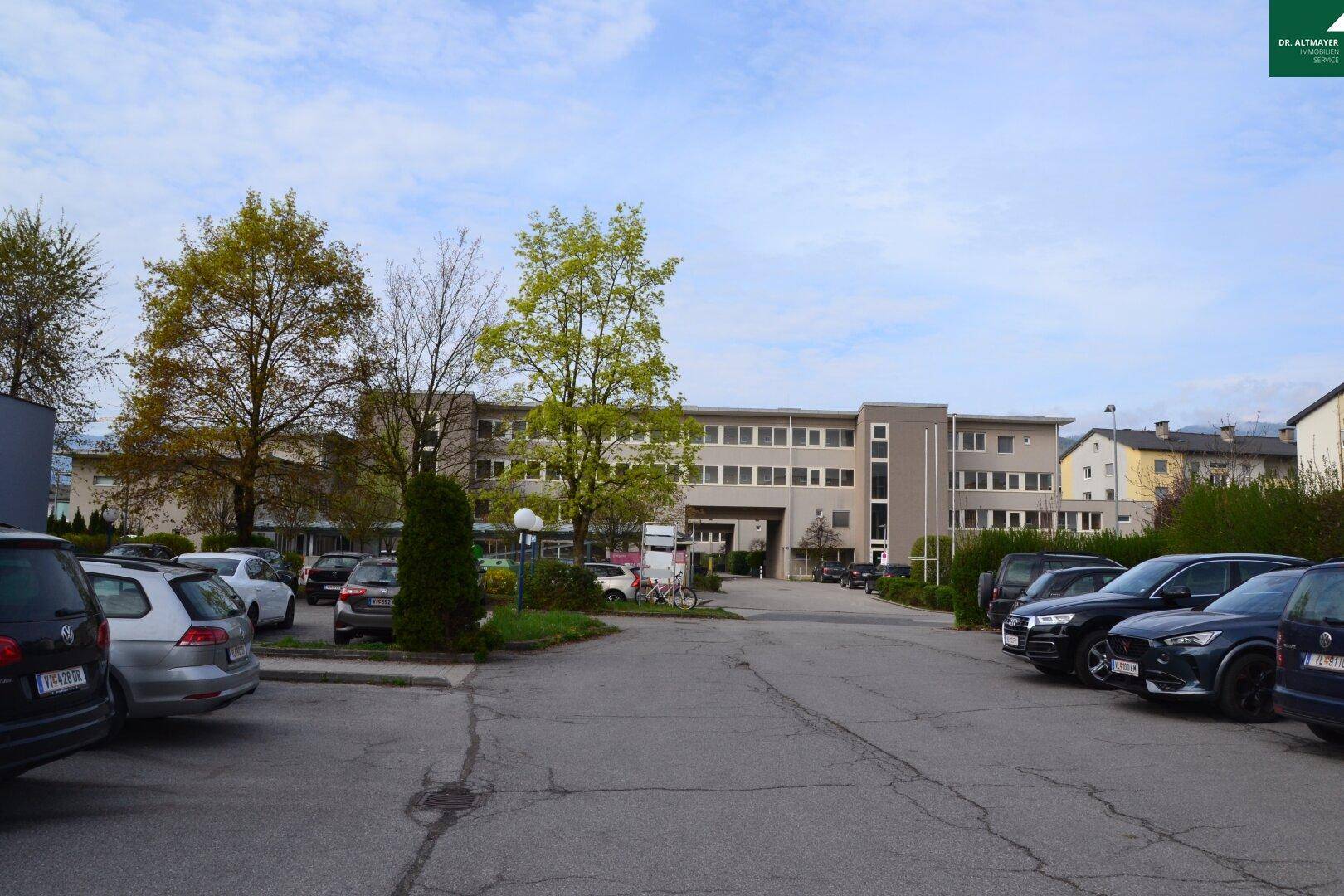 Weitläufiges Gebäude mit beiger Fassade und vielen Fenstern, davor ein großer Parkplatzbereich.