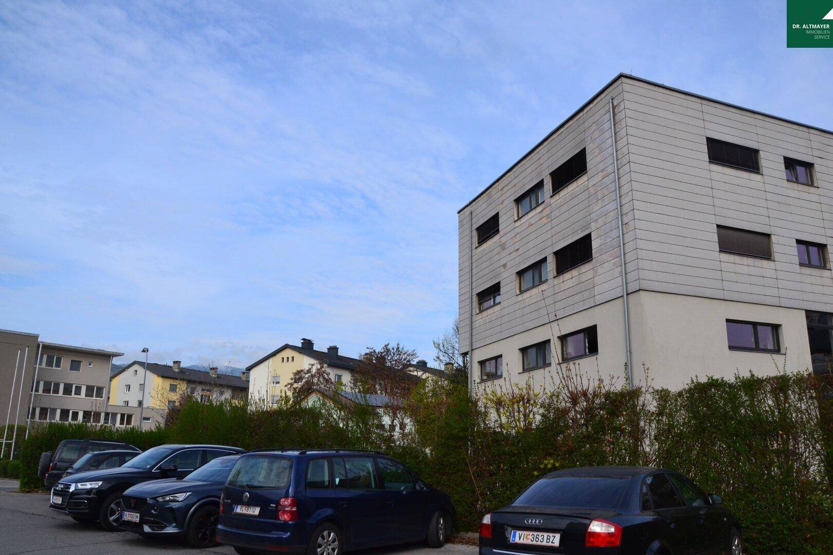 Modernes Gebäude mit grauer Paneelfassade, umgeben von einem Parkplatz und weiteren Gebäuden.