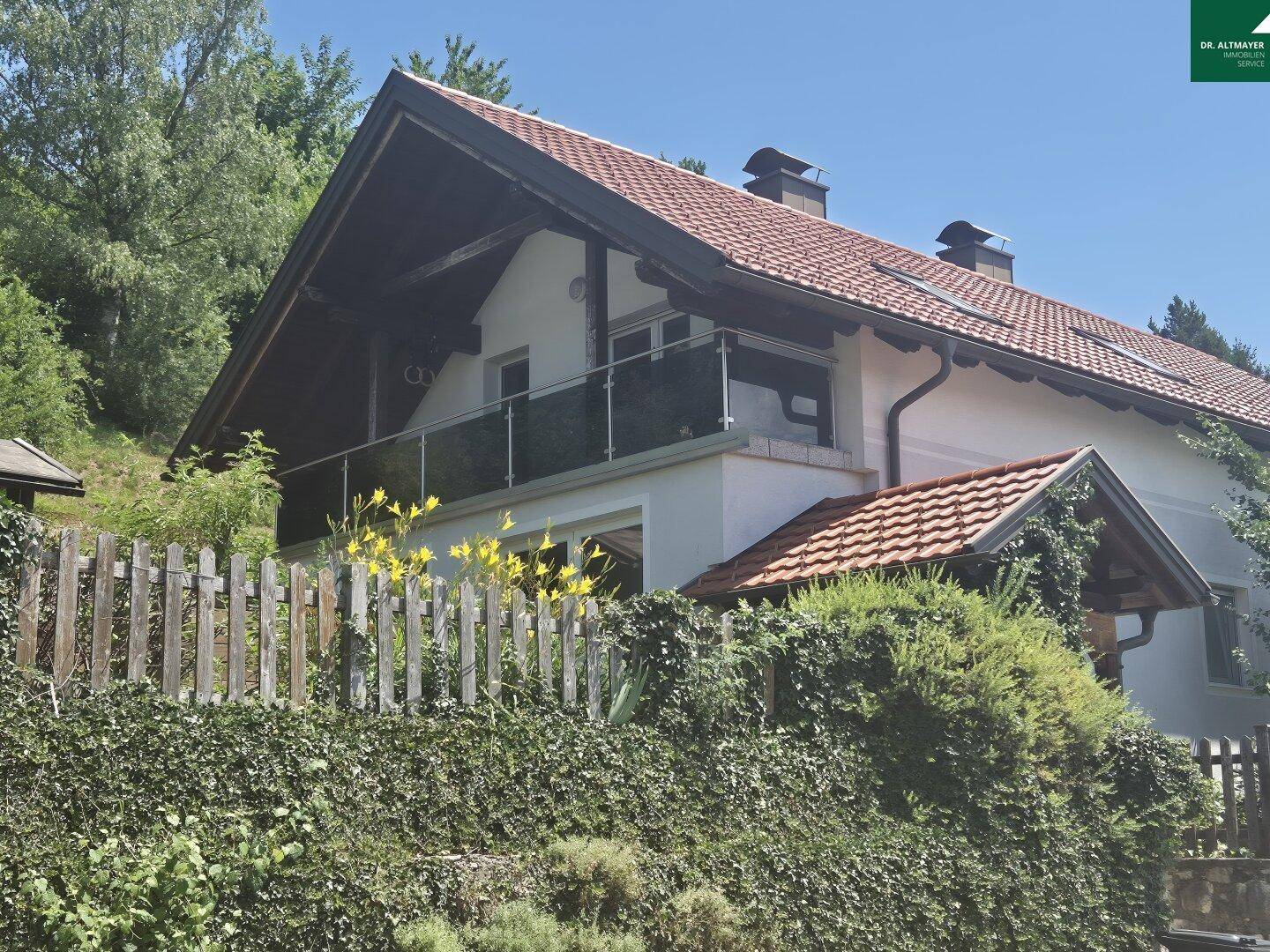 Seitenansicht des Hauses mit Satteldach, Balkon und üppiger grüner Umgebung.