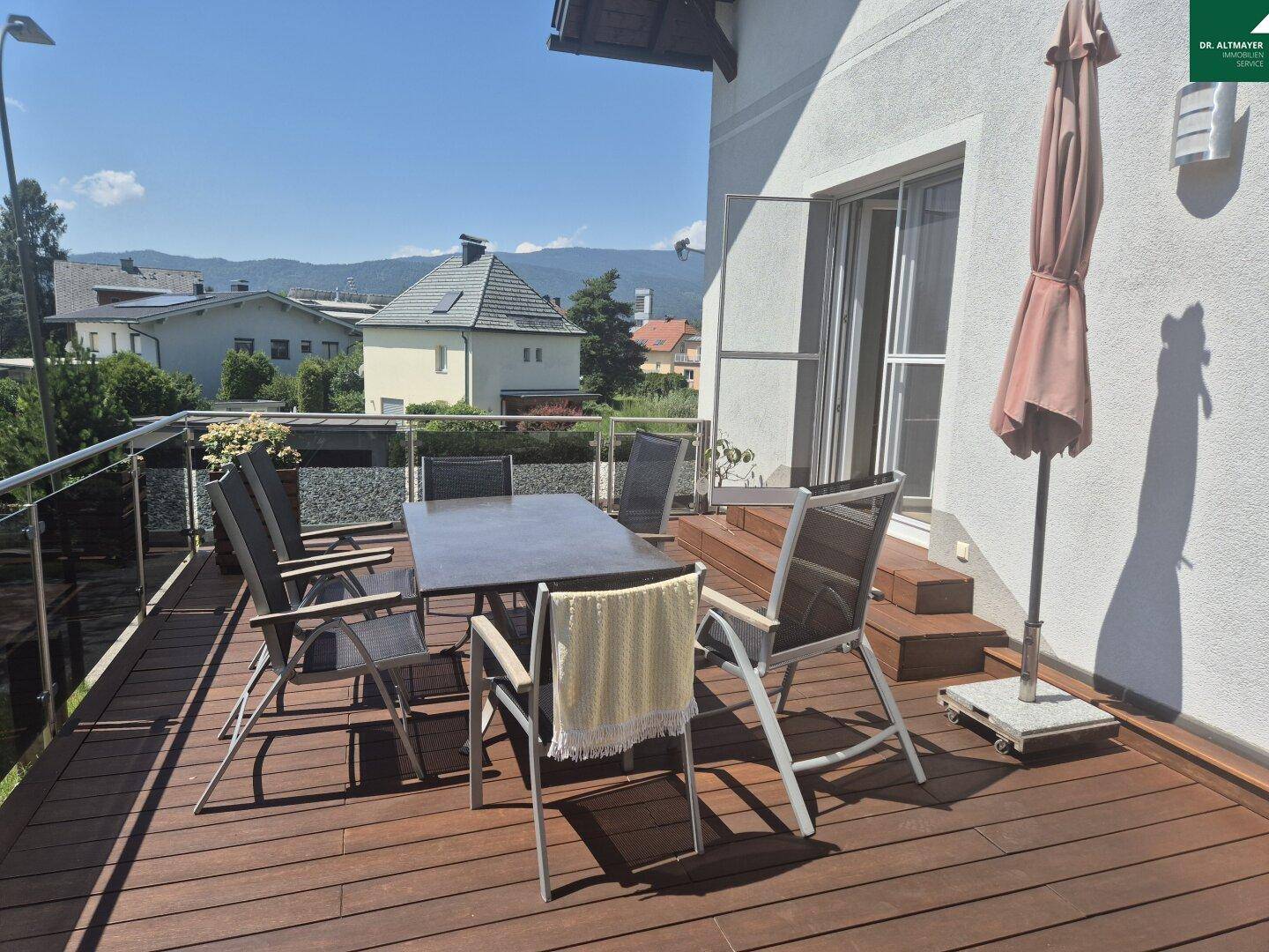 Geräumige Holzterrasse mit Esstisch, Sonnenschirm und Blick auf die Umgebung.