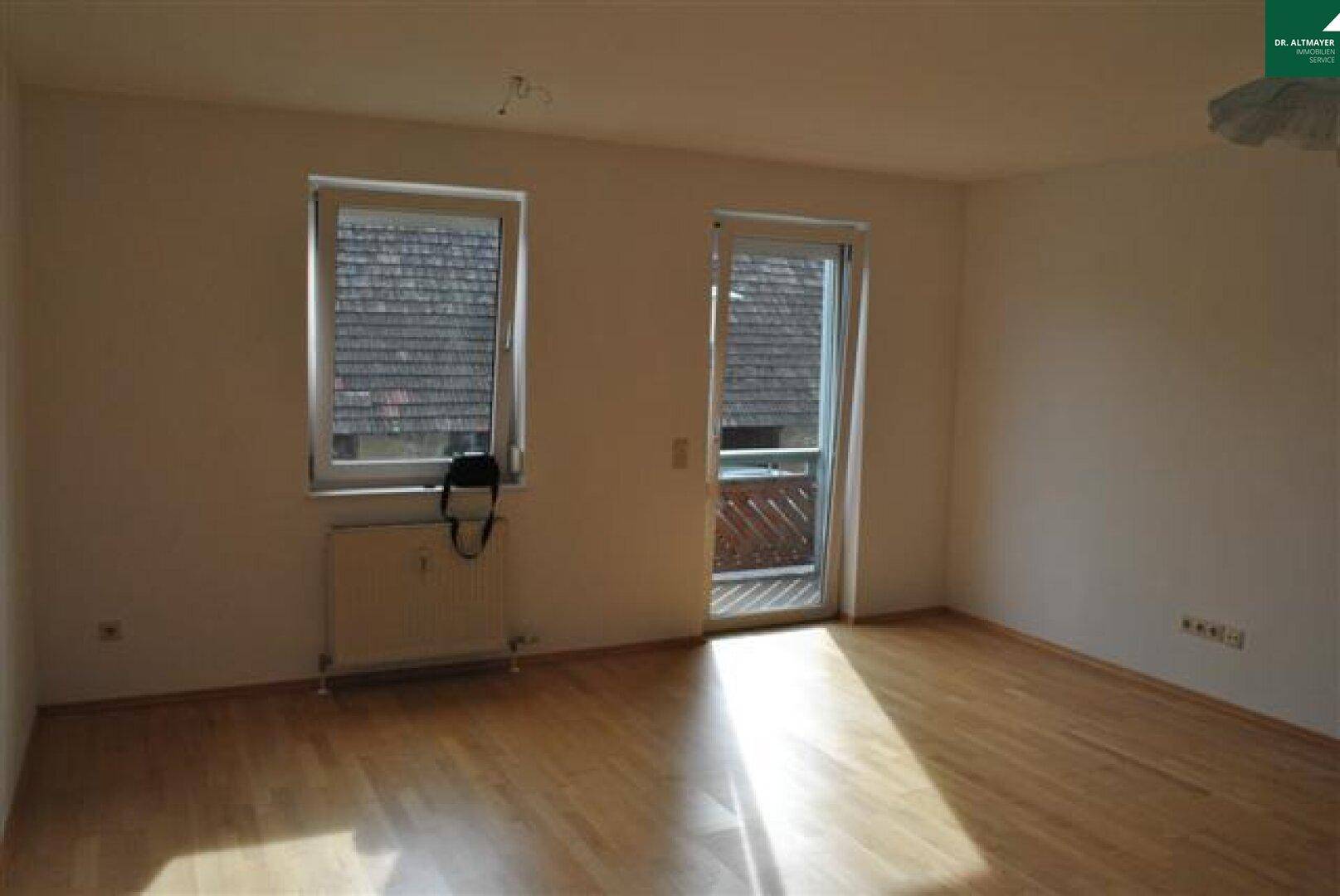 Leerer Raum mit hellem Parkettboden, Fenster und Zugang zu einem kleinen Balkon.