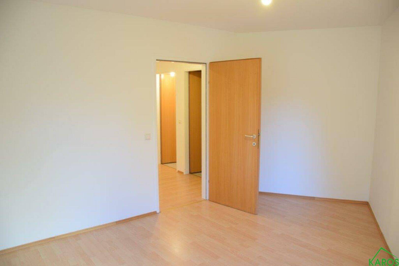 Leeres Zimmer mit Laminatboden und einer Holztür, die in einen Flur führt.