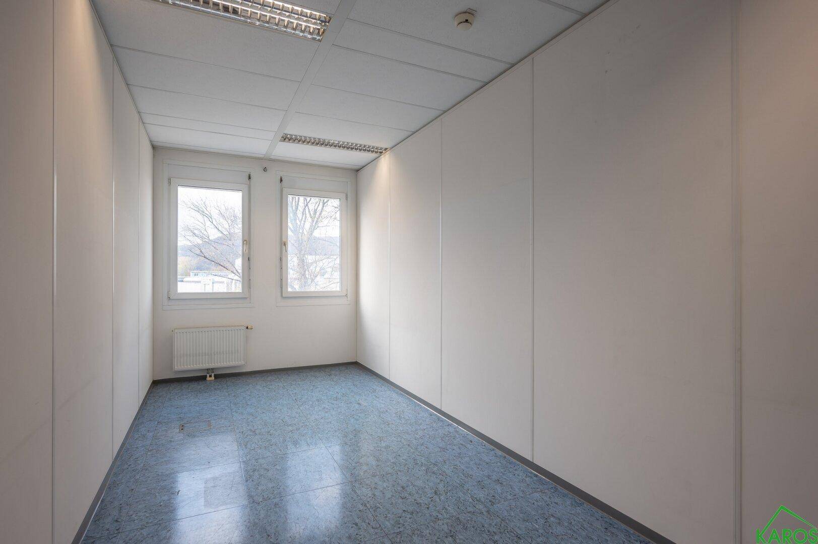 Kleines Büro mit zwei Fenstern, blauem Vinylboden und weißen Wänden, ideal für Einzelarbeit.