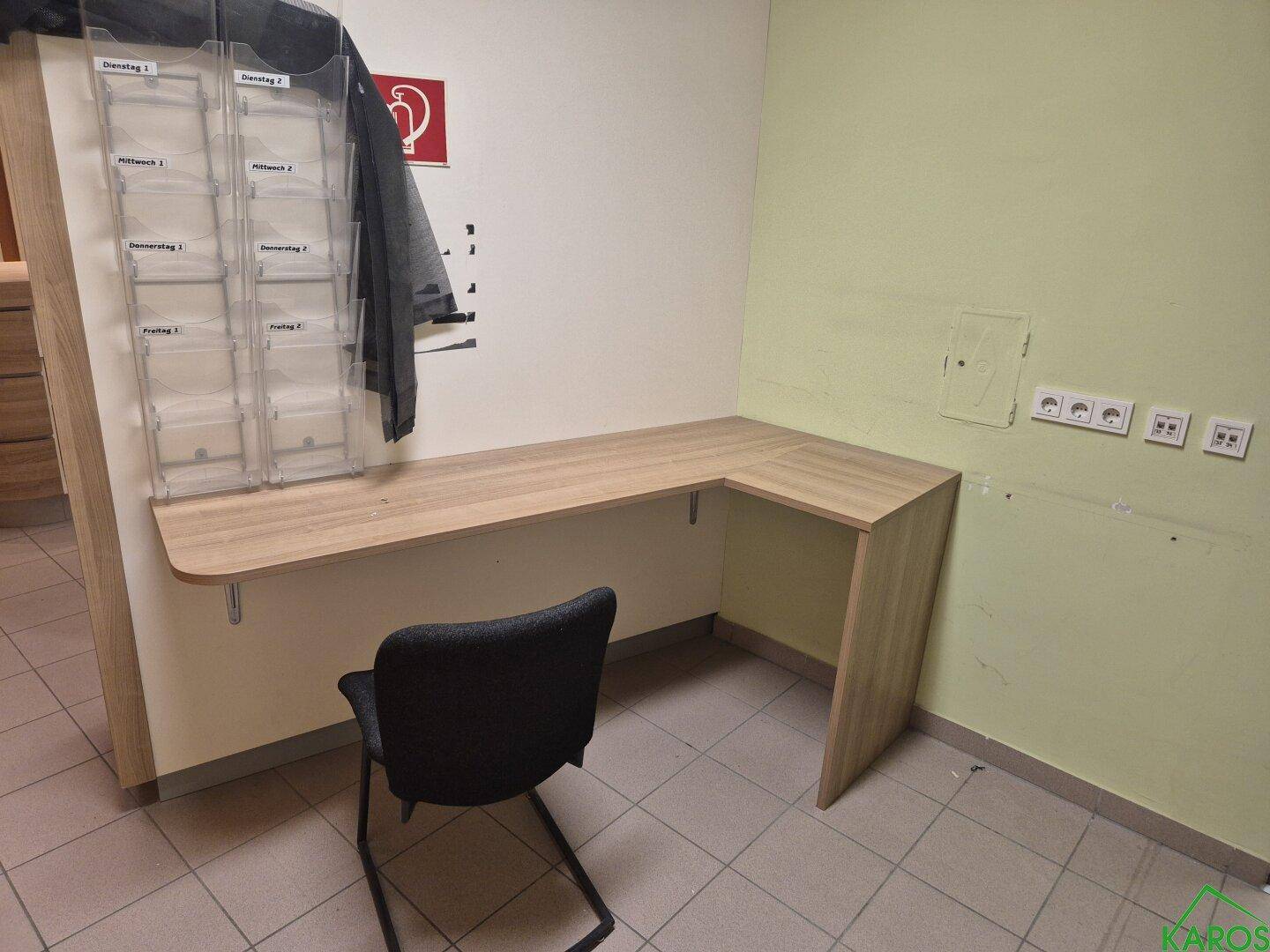 Kleiner Bürobereich mit Eckschreibtisch aus Holz und Fliesenboden, ausgestattet mit Steckdosen.