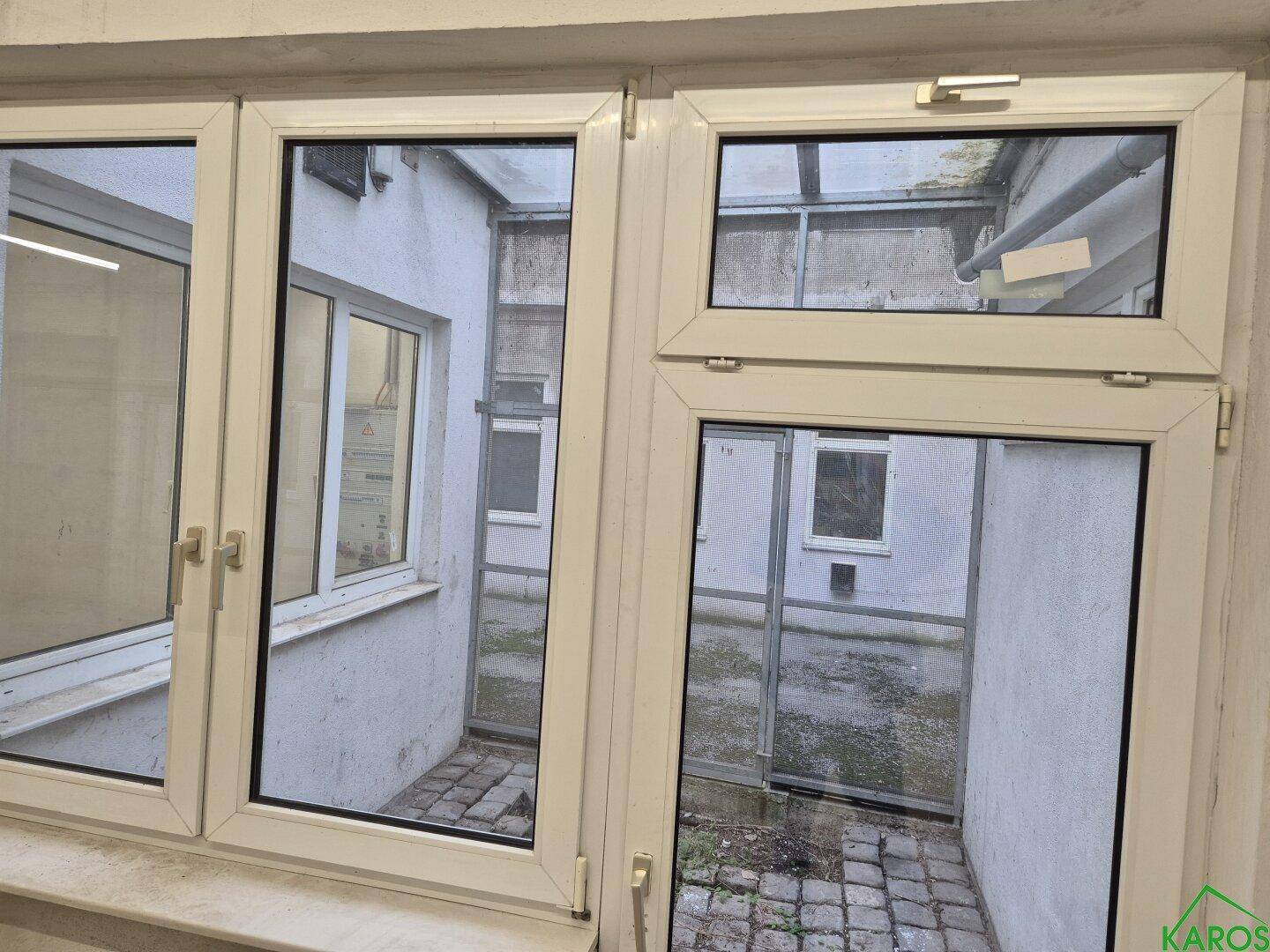 Fenster mit Blick auf einen kleinen, gepflasterten Innenhof mit weißen Wänden und Entwässerung.