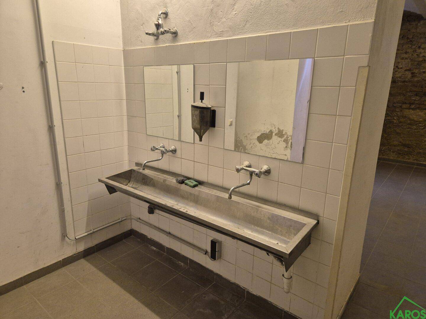 Industrieller Waschraum mit langem Trogwaschbecken, zwei Spiegeln und weiß gefliesten Wänden.