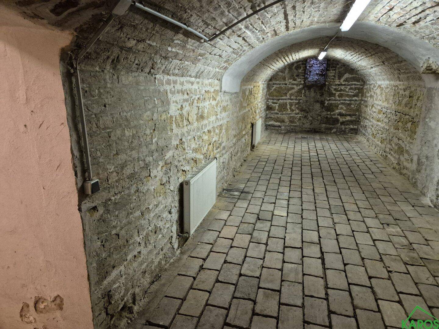 Gewölbekeller mit Natursteinwänden und gepflastertem Boden, beleuchtet von Leuchtstoffröhren und einem kleinen Fenster.