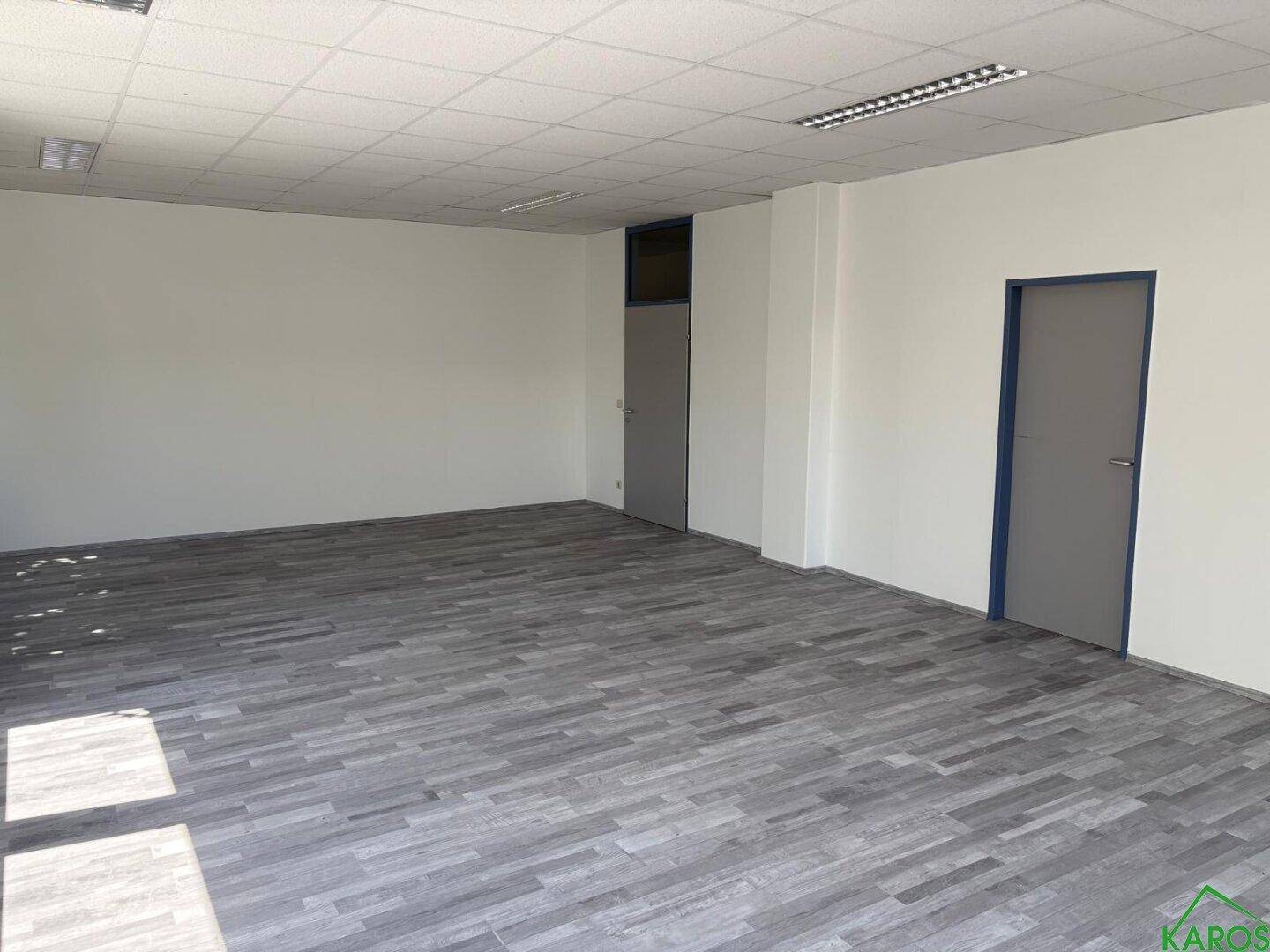 Leerer Büroraum mit grauem Laminatboden, weißen Wänden und zwei Türen.