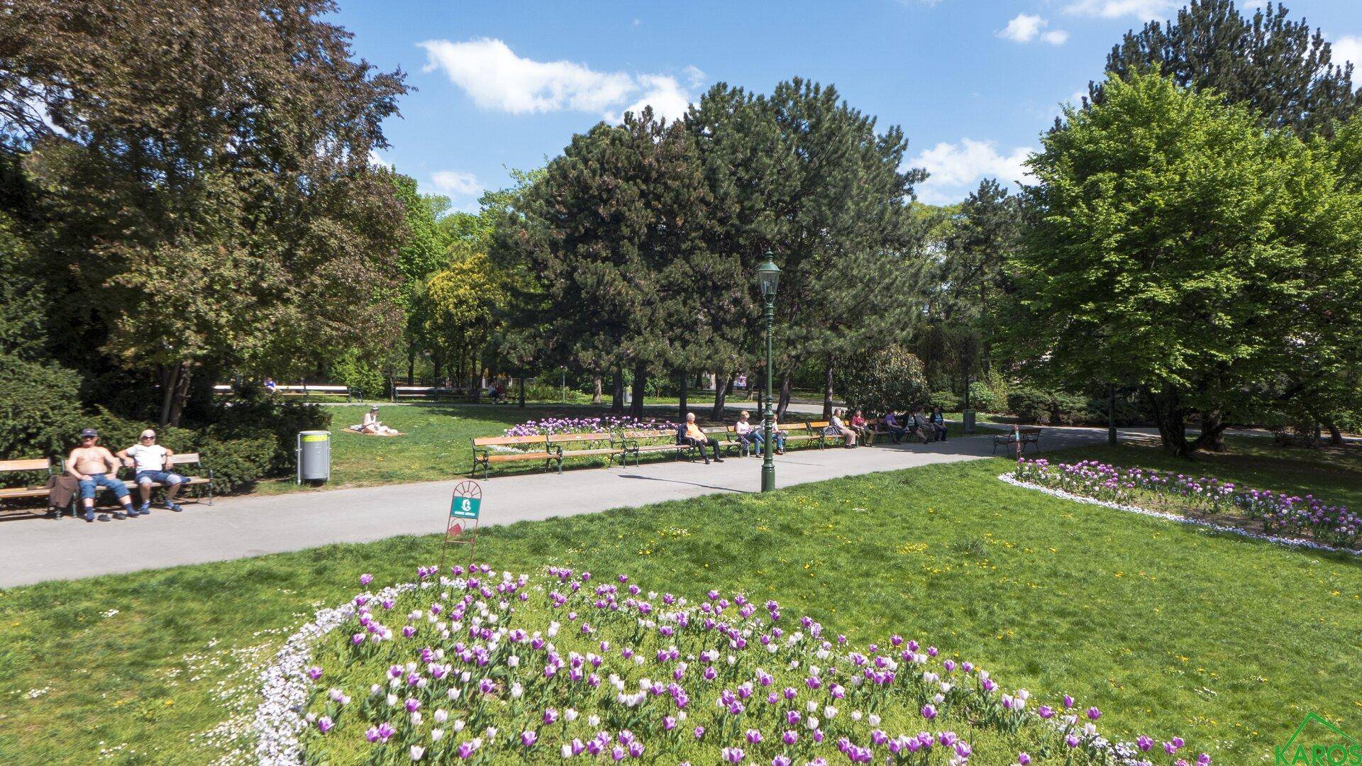 Gepflegter Park mit Blumenbeeten, Wegen und Sitzbänken unter sonnigem Himmel.