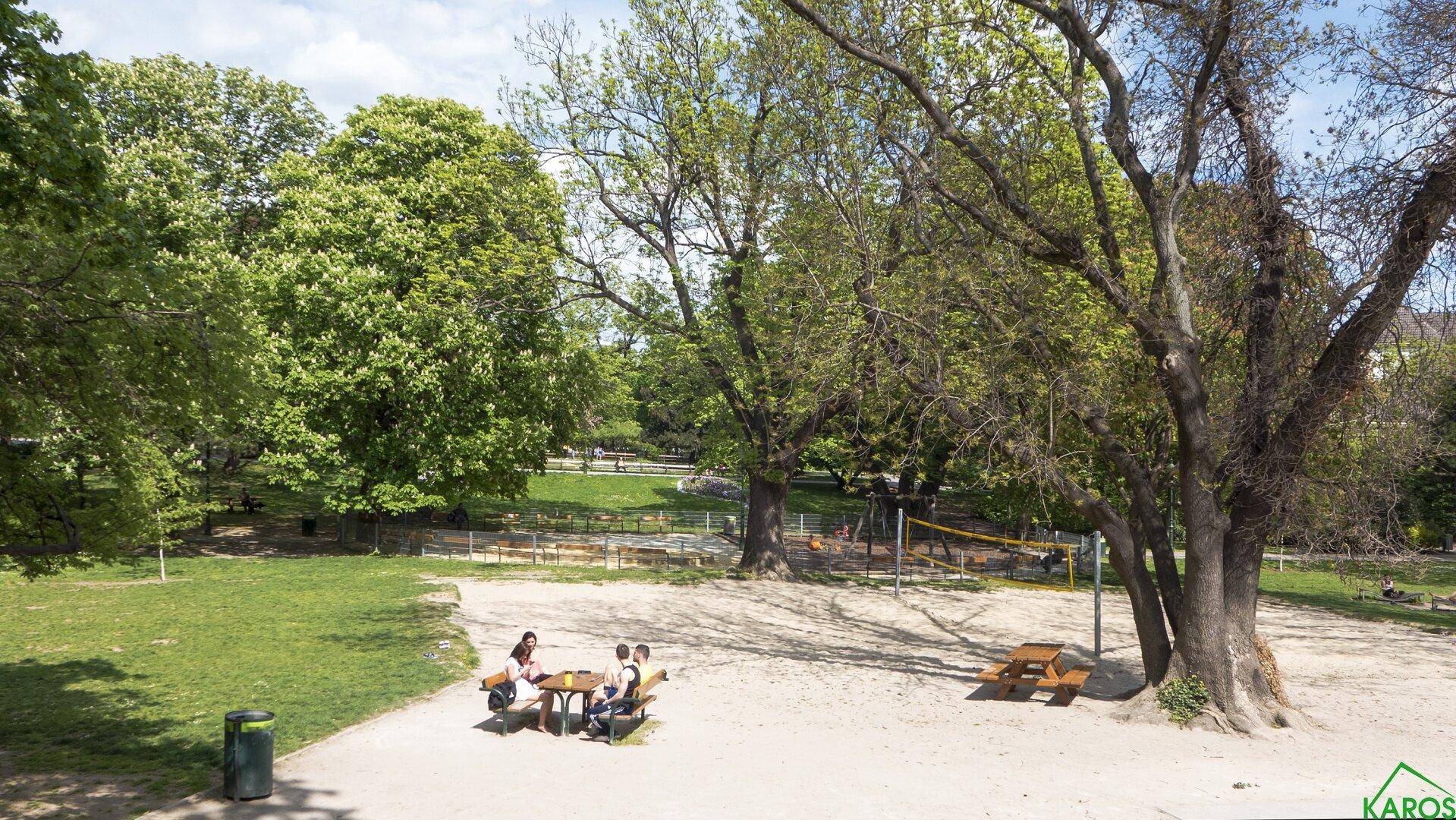 Sandiger Spielbereich in einem Park mit Bäumen und Sitzgelegenheiten für Erholung.