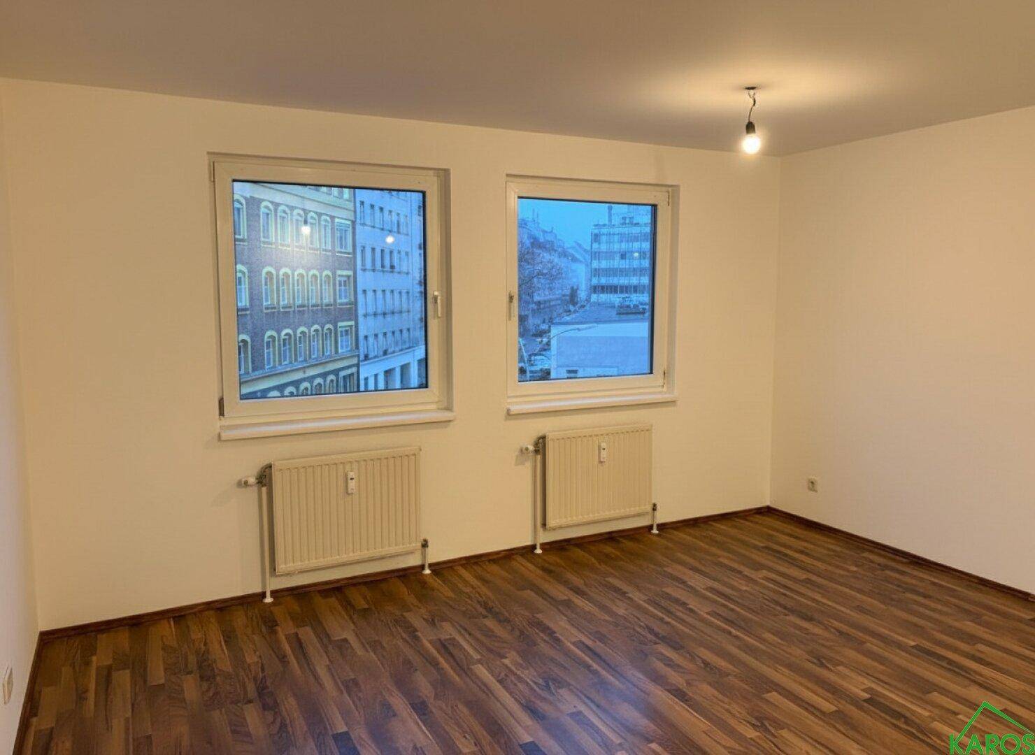 Geräumiges, unmöbliertes Wohnzimmer mit Laminatboden und zwei großen Fenstern.