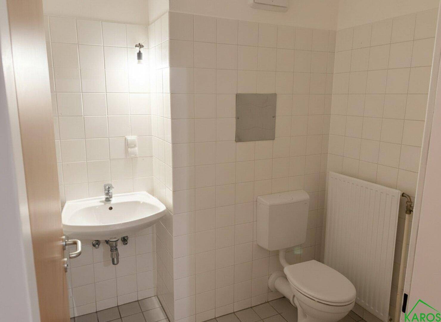 Separates WC mit Waschbecken und Heizkörper, vollständig gefliest in Weiß.