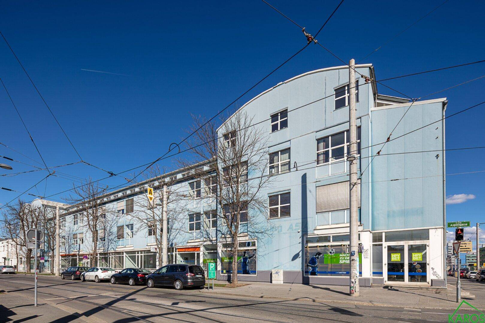 Gebäudeansicht von der Straßenecke mit blauer Fassade, vielen Fenstern und Geschäften im Erdgeschoss.