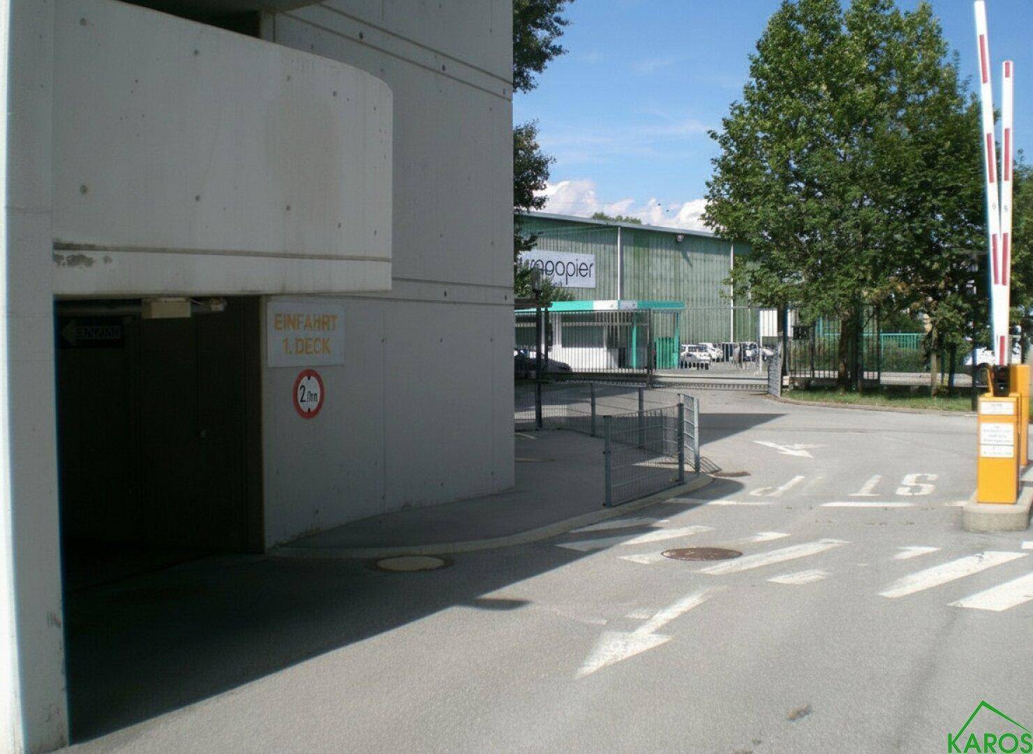Einfahrt zu einem Parkhaus mit Schranke und deutlicher Beschilderung, umgeben von Bäumen und einem weiteren Gebäude.