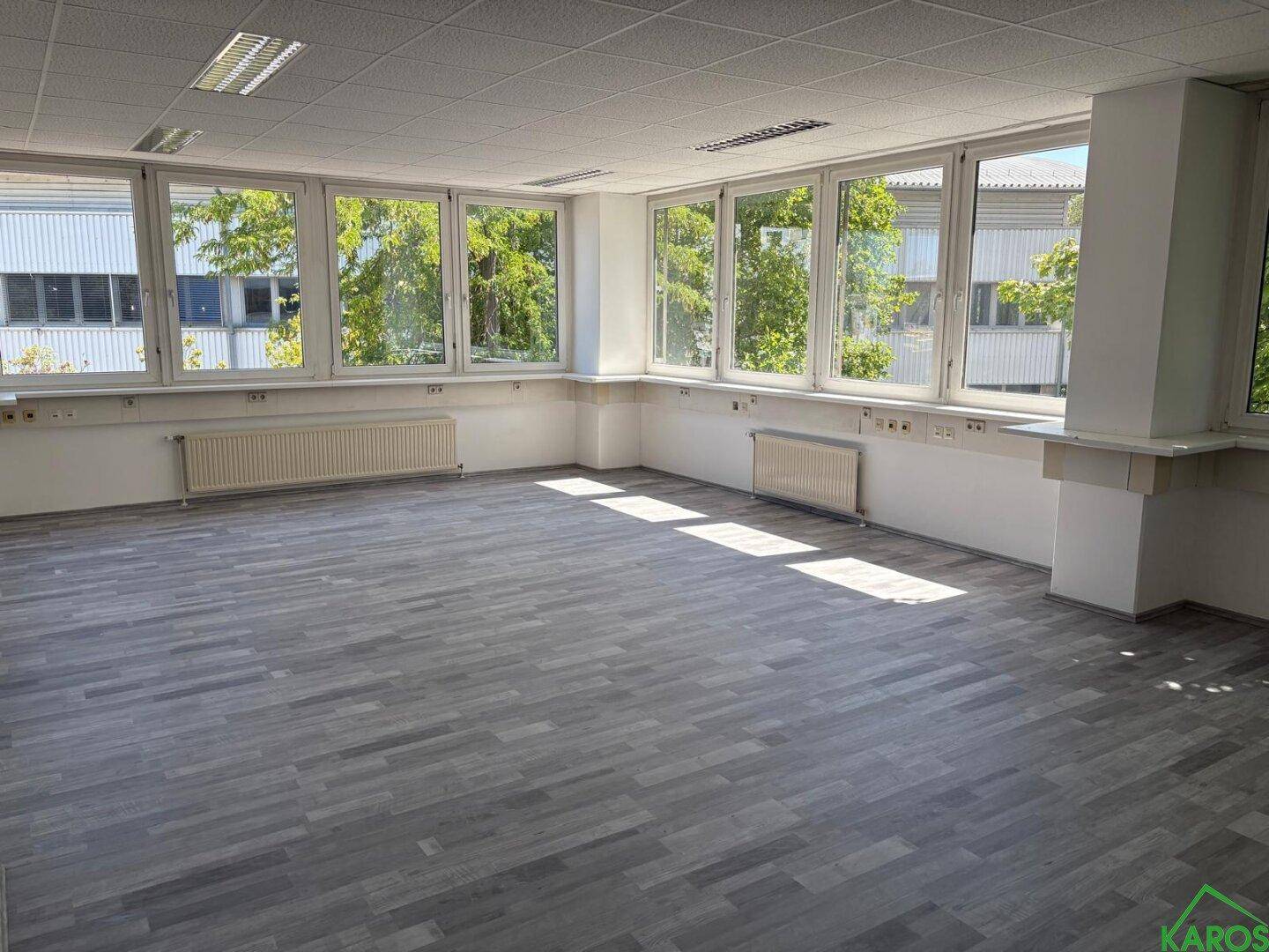 Leerer, heller Büroraum mit großen Fenstern, Laminatboden und Heizkörpern unter den Fenstern.