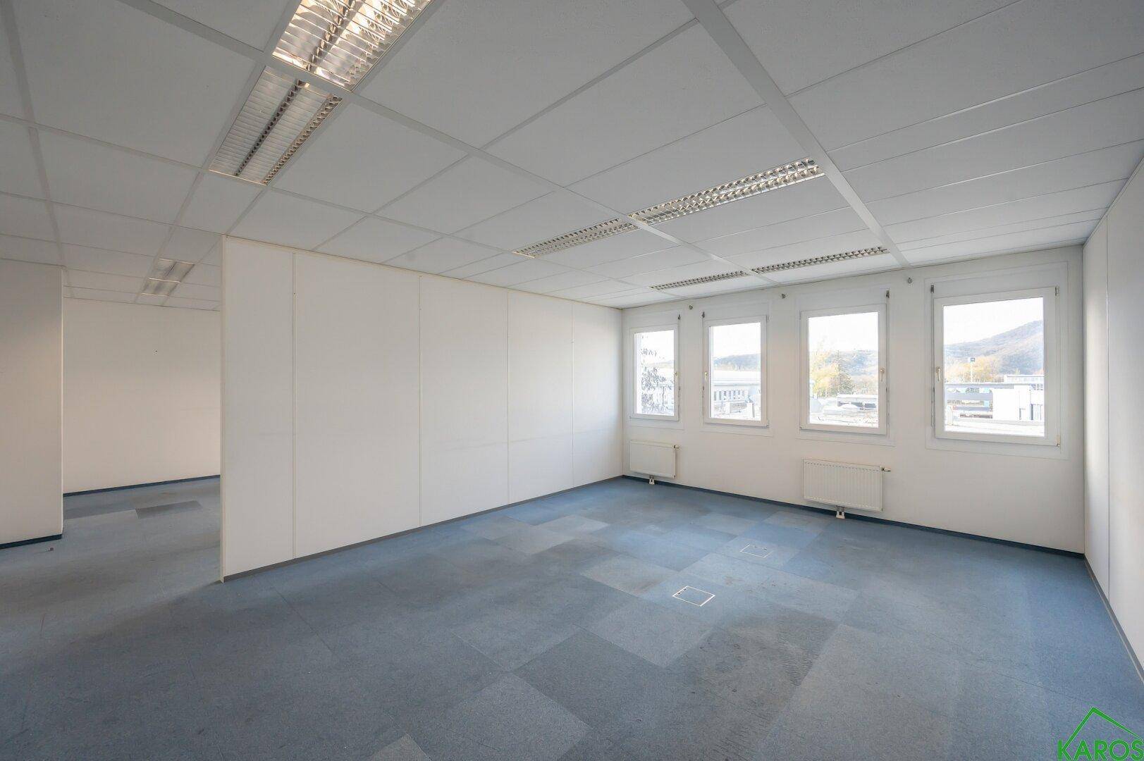 Helles, leeres Büro mit blauer Teppichboden und vier Fenstern, das einen Blick in einen angrenzenden Raum bietet.